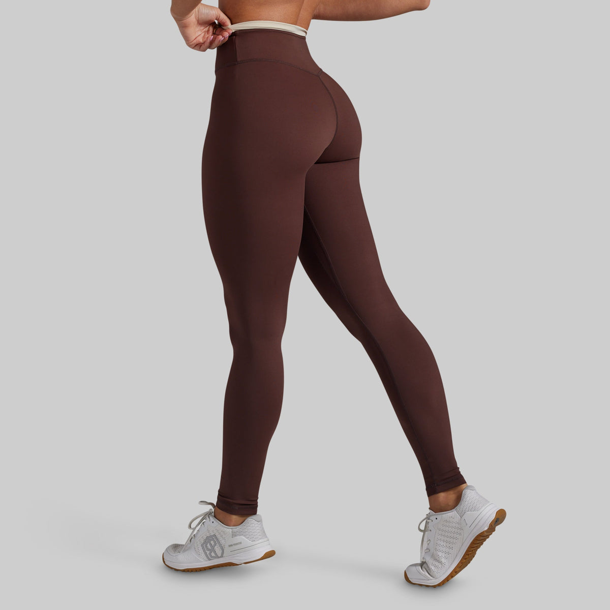 A model wearing high-waisted, form-fitting leggings in a dark brown color, shown from a rear three-quarter angle against a solid light gray background.