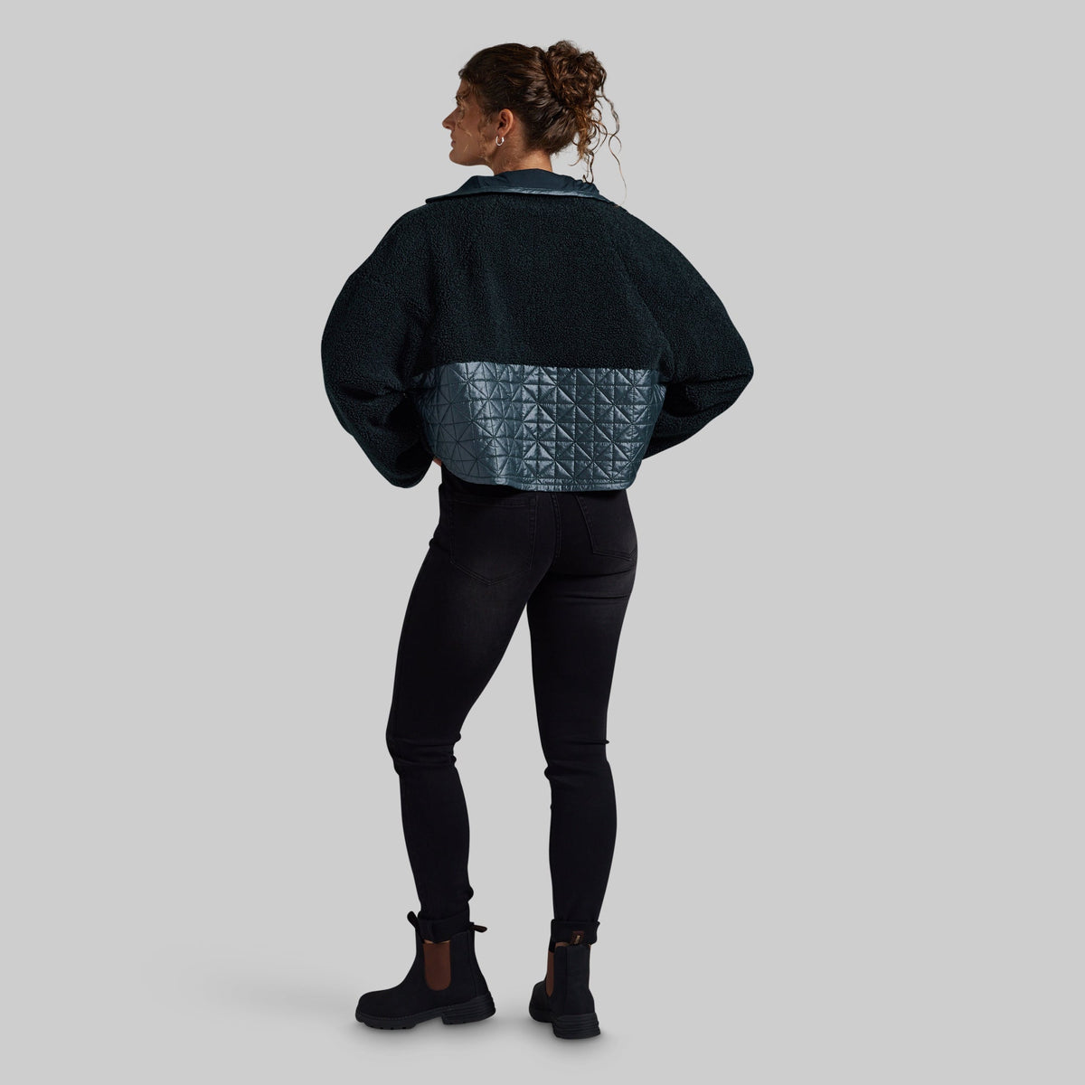 A woman is shown from the back wearing a dark teal cropped jacket with a textured geometric quilted panel across the lower back, paired with black leggings and black ankle boots, against a solid gray background.