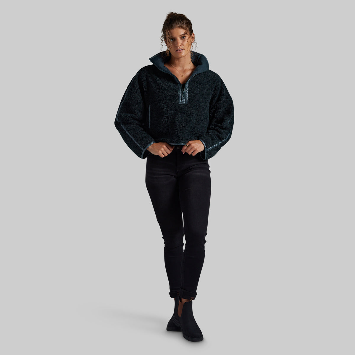 A woman modeling a dark teal fleece pullover with a half-zip neckline and ribbed cuffs, paired with black pants and black ankle boots, against a solid light gray background.