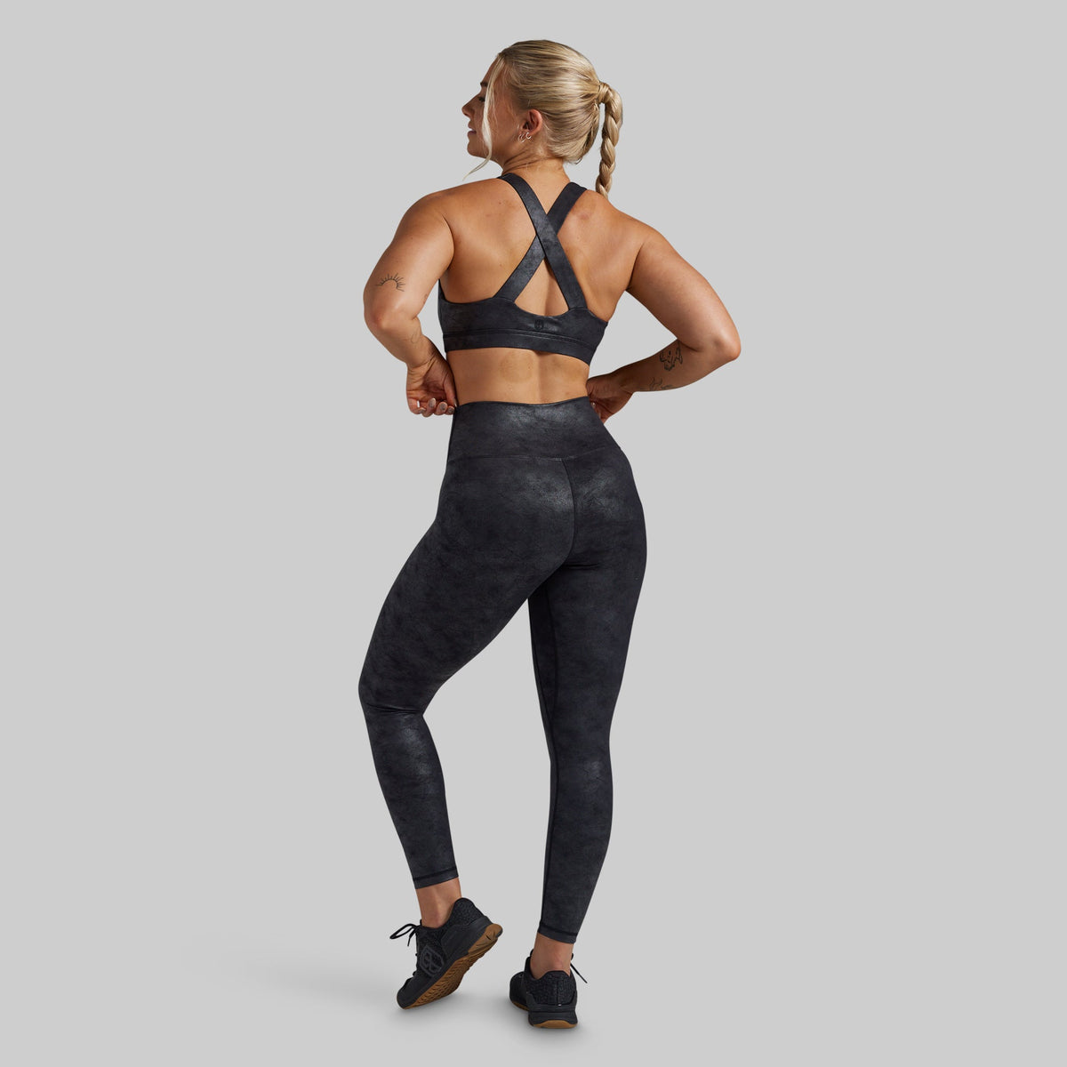 A woman shown from the back wearing a matching dark gray and black athletic set, featuring a sports bra with crisscross straps and high-waisted leggings with a subtle mottled pattern, set against a solid gray background.