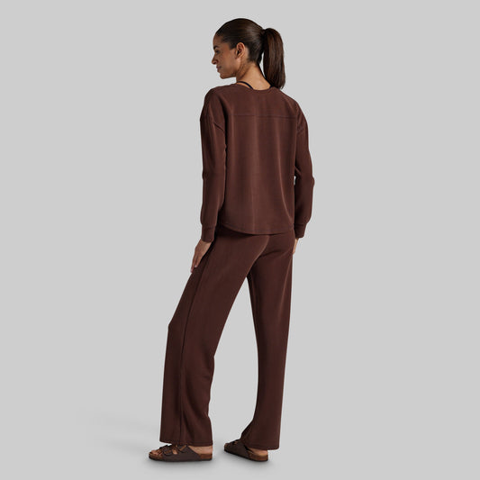 A woman wearing a matching dark brown lounge set consisting of a long-sleeve top and wide-leg pants, shown from a rear three-quarter angle against a solid gray background.