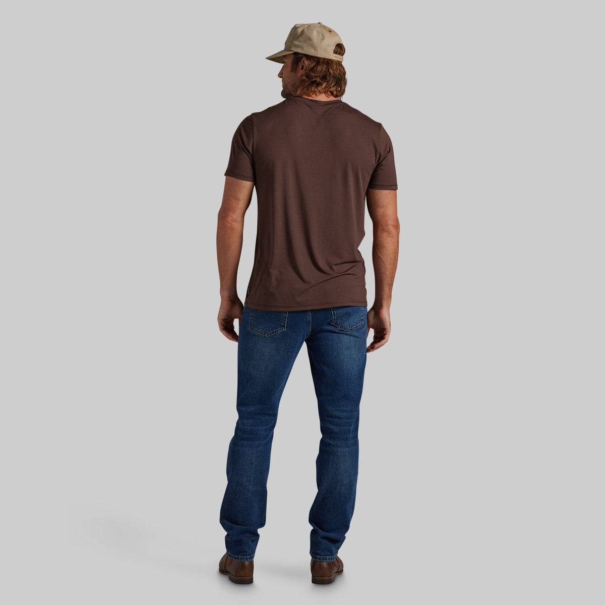 A man viewed from behind wearing a dark brown short-sleeve t-shirt, blue jeans, and a beige baseball cap, standing against a solid gray background.