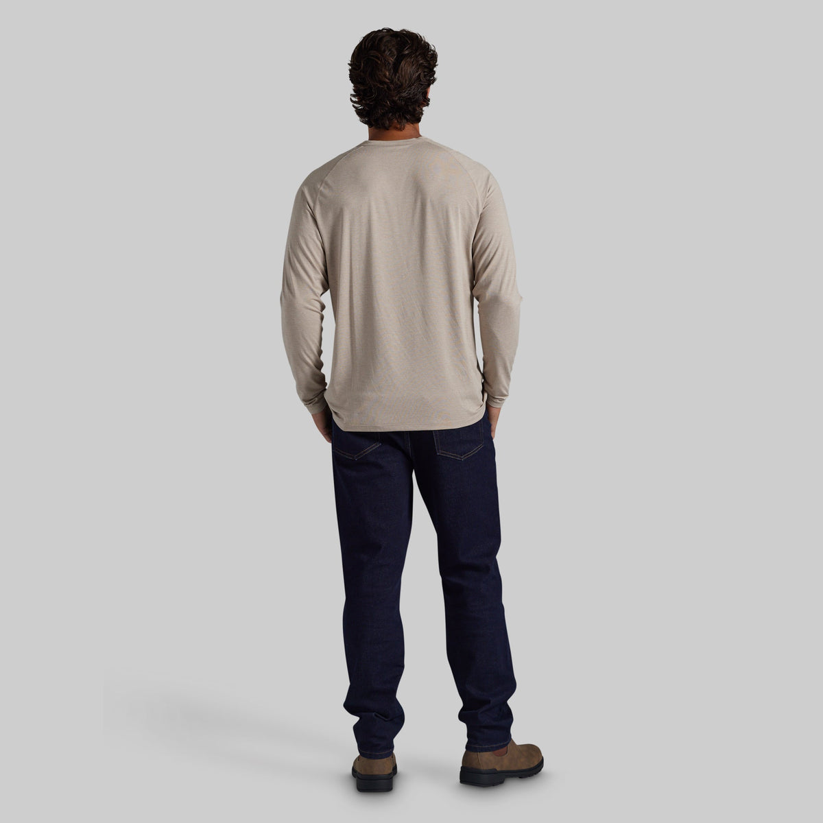 A man viewed from behind wearing a light beige long-sleeve crewneck shirt, dark navy blue jeans, and brown boots, standing against a solid light gray background.