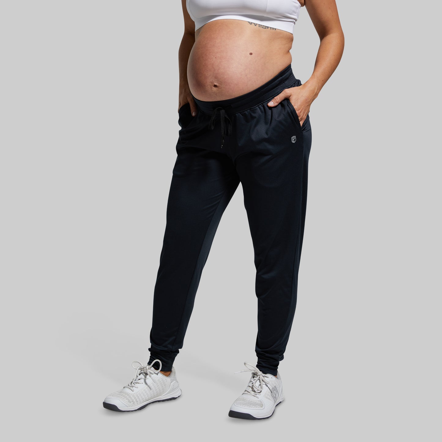 Pregnant person wearing black jogger pants and white sneakers on a plain background