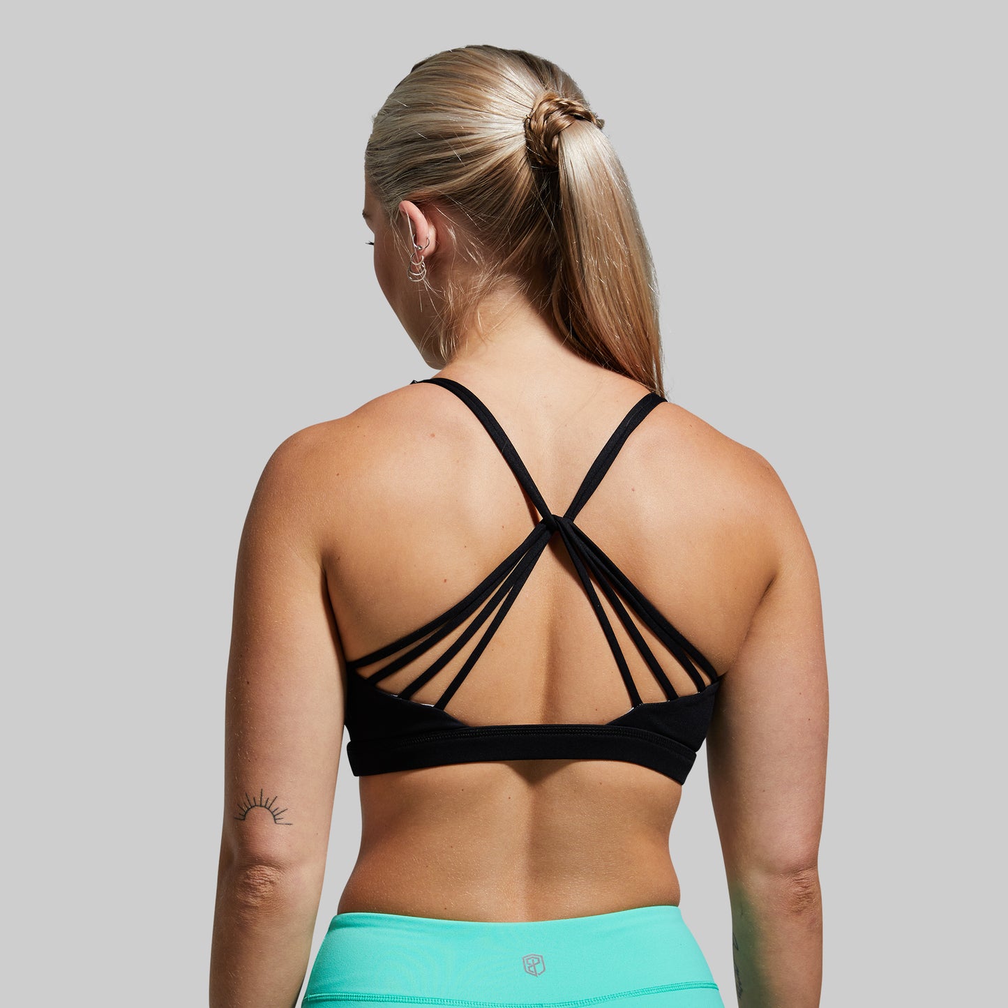 Woman wearing a black strappy sports bra and teal shorts on a gray background