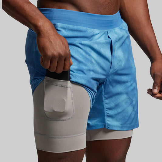 Blue athletic shorts with a gray pocket worn by a person on a gray background.