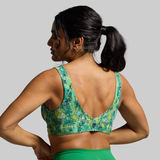 A woman wearing a green and multicolor floral sports bra with over-the-shoulder back straps, facing away from the camera.