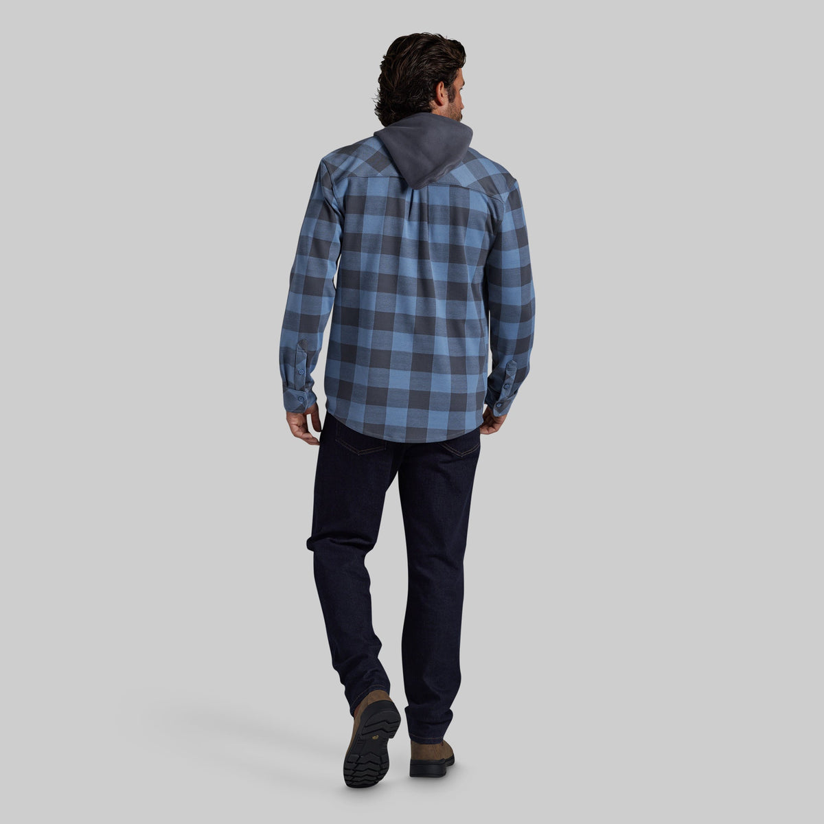 A man viewed from behind wearing a blue and gray plaid hooded flannel, dark navy pants, and brown shoes, standing against a solid light gray studio background.