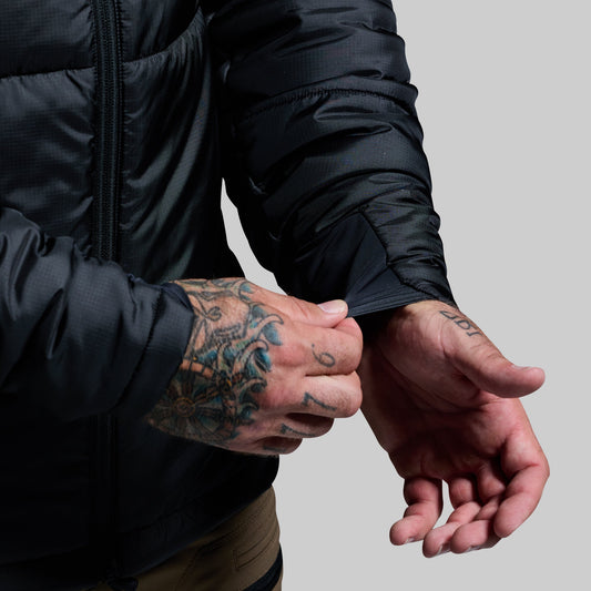 Person wearing a black jacket with visible tattoos on arms against a gray background