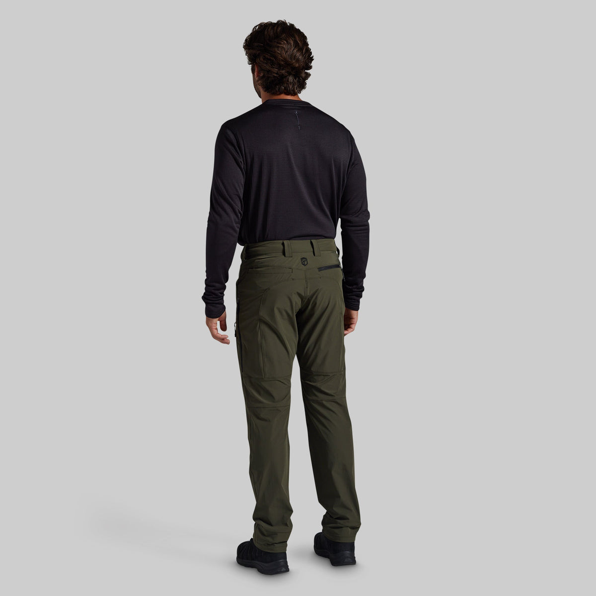 A man viewed from behind wearing a black long-sleeve shirt and olive green cargo pants against a solid gray background.