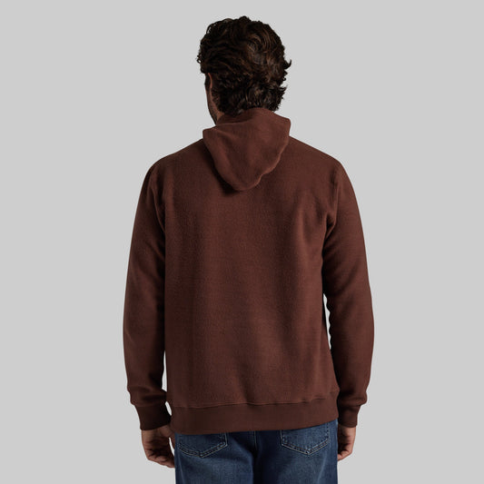A man viewed from behind wearing a dark brown pullover hoodie and blue jeans, set against a solid light gray background.