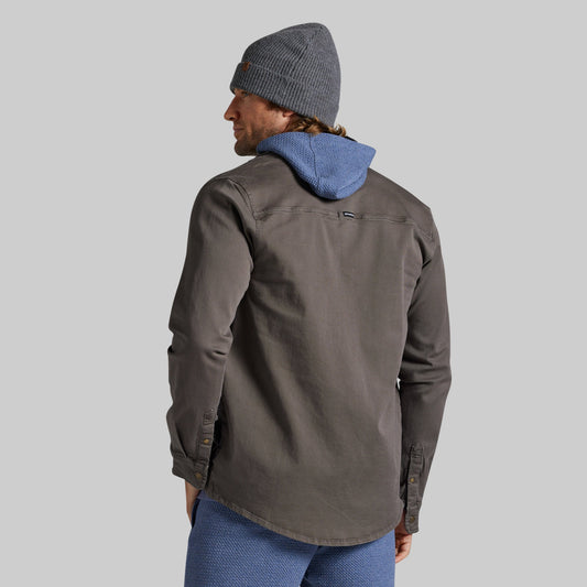 A man shown from the back wearing a dark gray overshirt, a gray beanie, and blue joggers against a solid gray background.