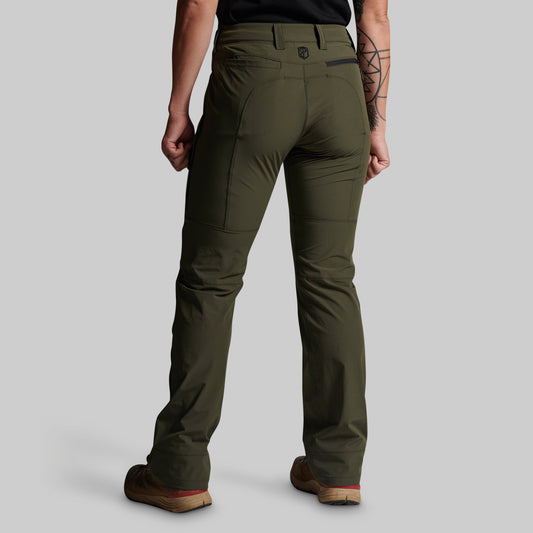 A person shown from the back wearing olive green technical pants, a black shirt, and brown shoes against a light gray studio background.