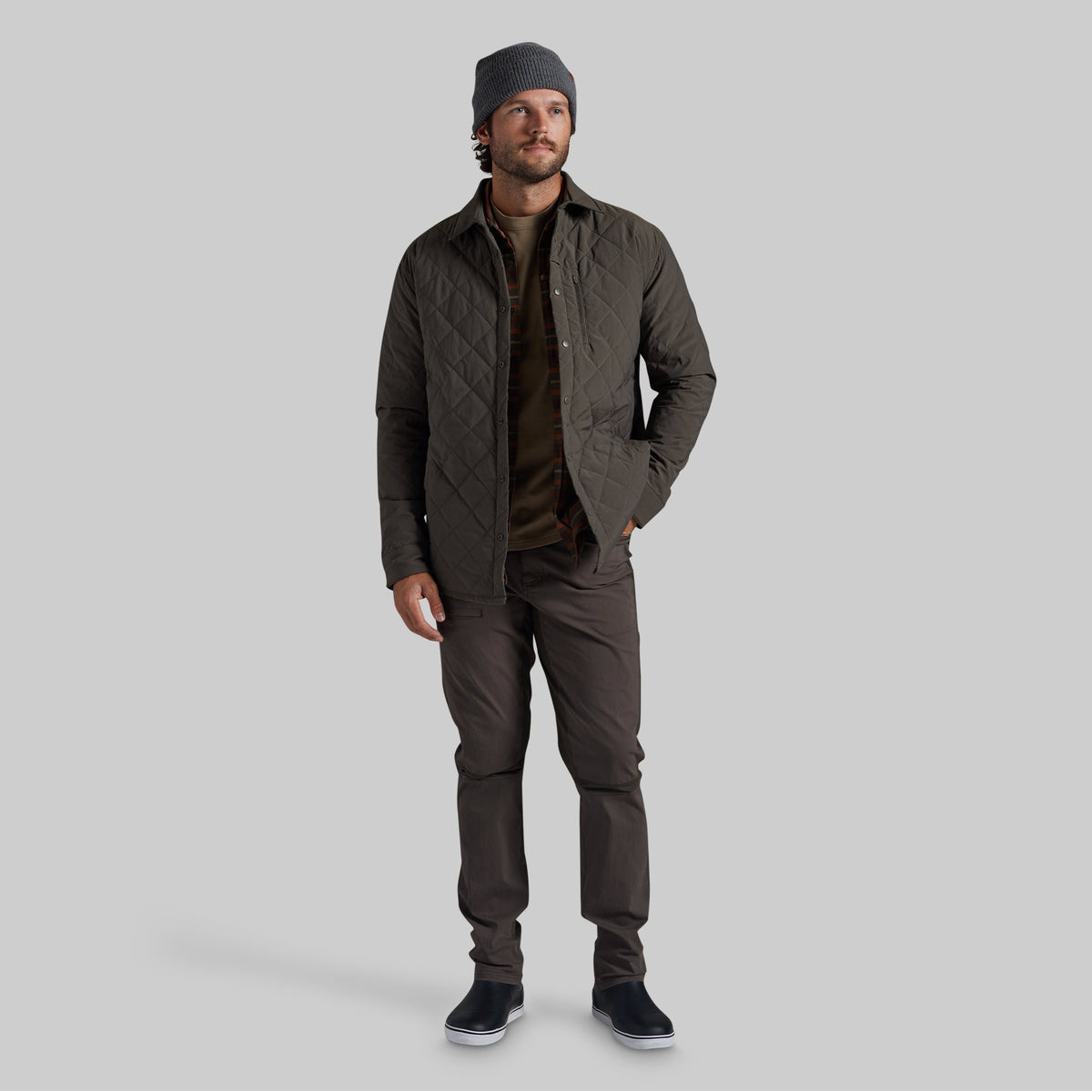 A man wearing a dark olive green quilted jacket, brown shirt, dark brown pants, a gray beanie, and black slip-on shoes, standing against a solid gray studio background.