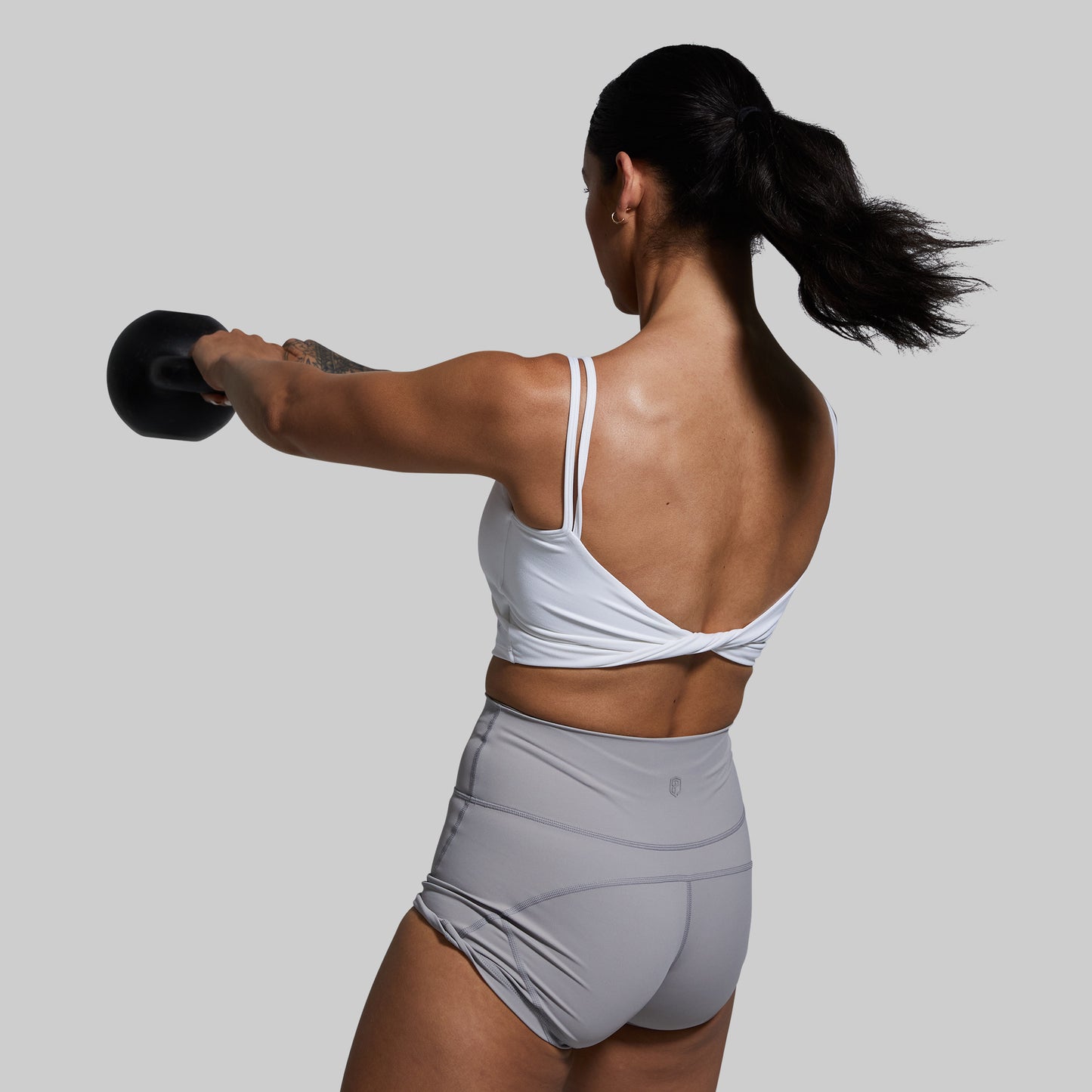Woman exercising with a kettlebell wearing a white sports bra on a plain background
