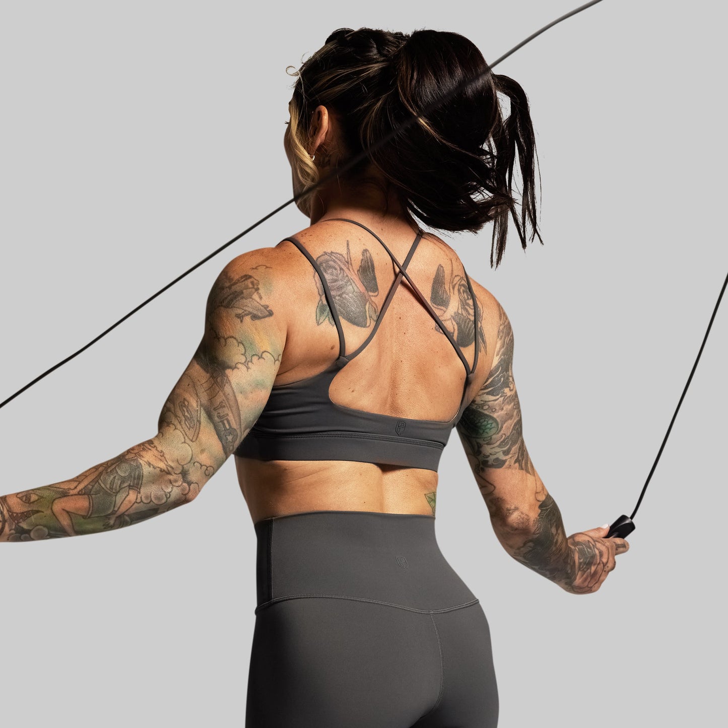 Person in gray athletic wear with tattoos holding a jump rope on a gray background
