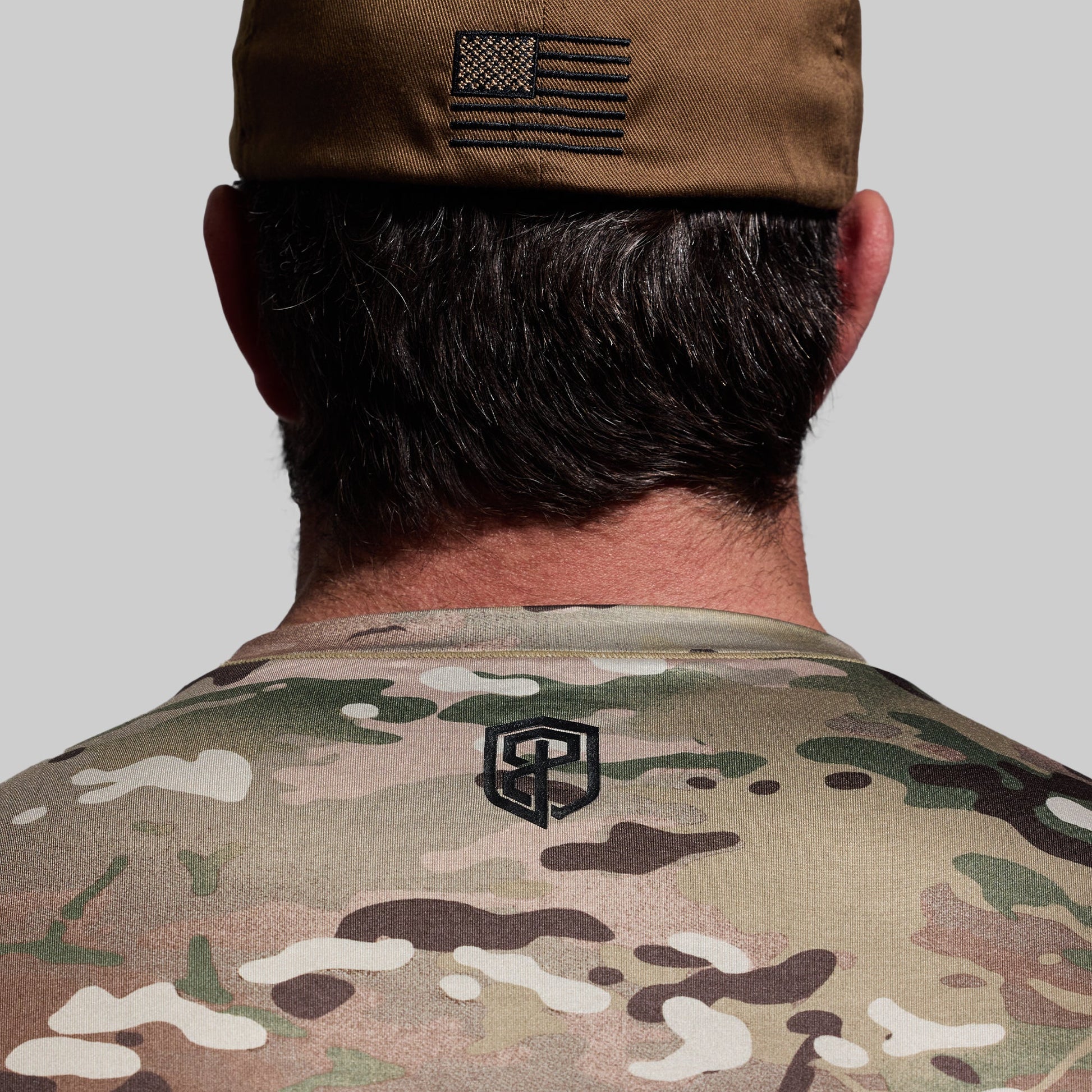 Person wearing a camouflage uniform with a cap featuring an American flag patch on a plain background