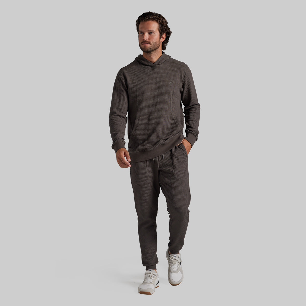 A man modeling a dark grey matching hoodie and sweatpants set, paired with white sneakers, against a solid light gray background.