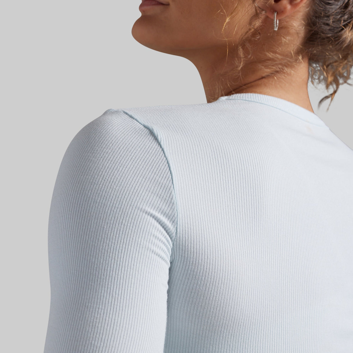 Close-up of a woman wearing a light icy blue ribbed long-sleeve top, showing the back and shoulder detail against a neutral gray background.
