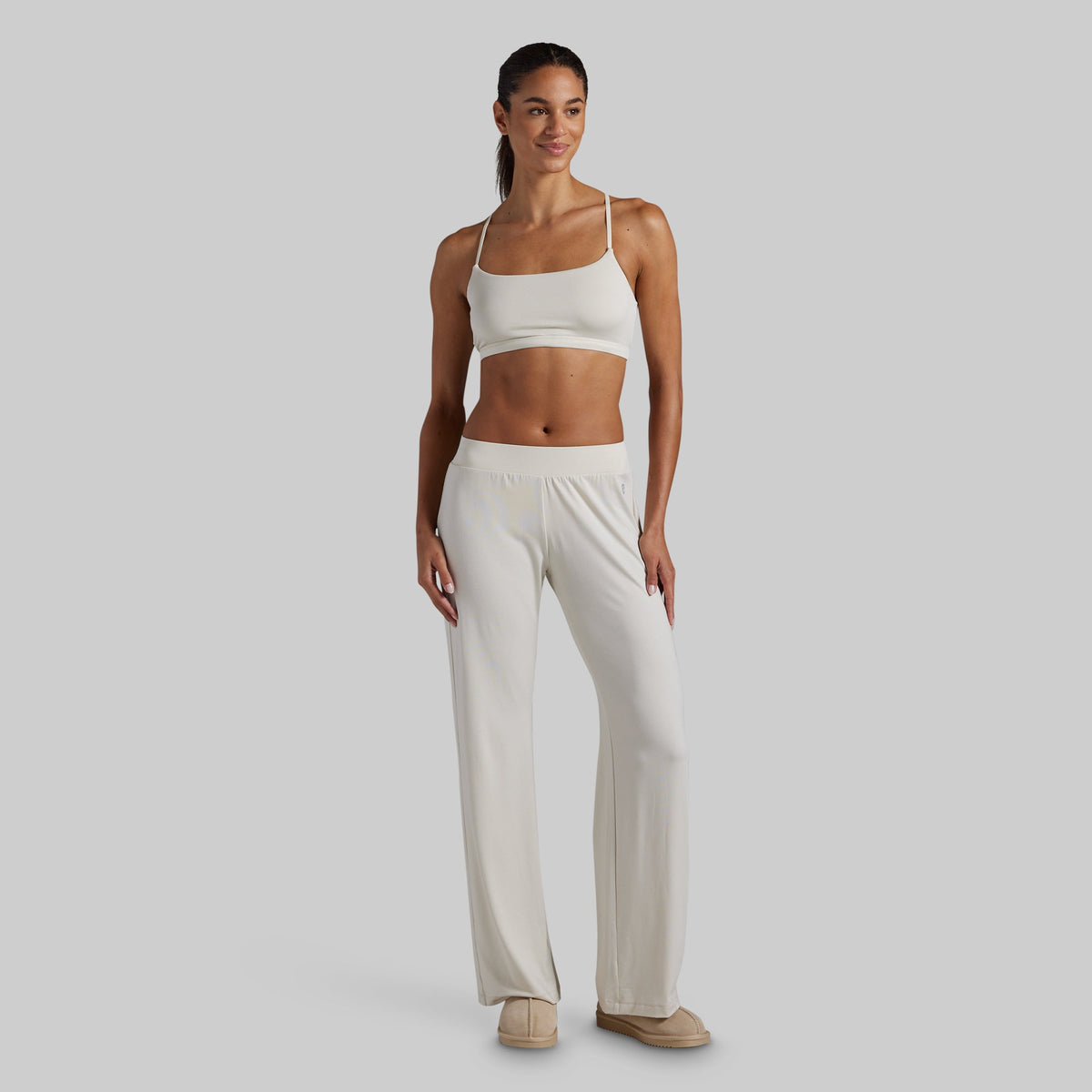 A woman modeling a matching two-piece lounge set consisting of a white sports bra and white wide-leg pants, standing against a solid light gray background.