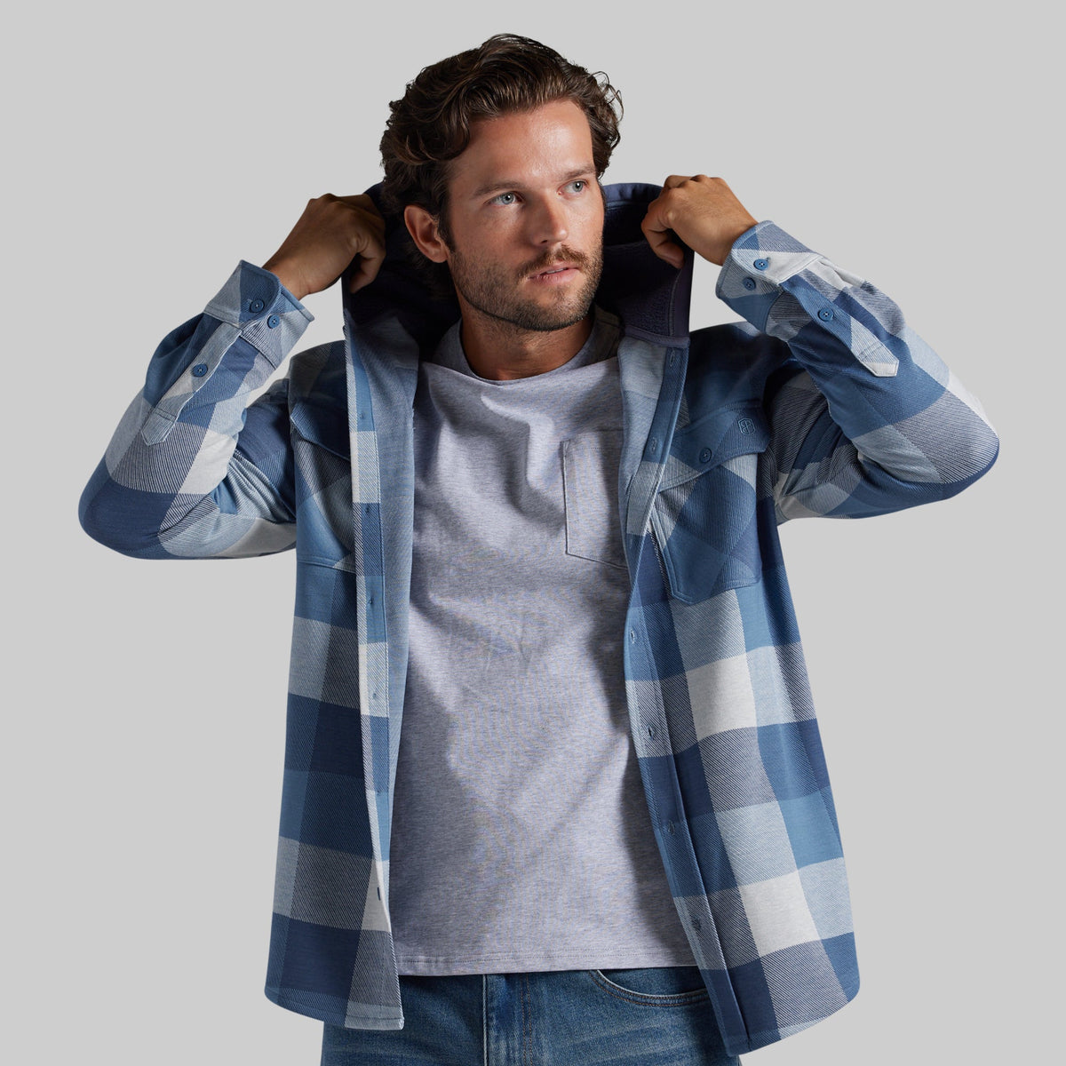 A man wearing a blue and white plaid overshirt over a light gray t-shirt and blue jeans, adjusting the hood of the flannel against a solid gray background.