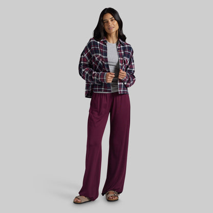 A woman modeling a two-piece lounge set consisting of a dark red and navy plaid flannel shirt and deep burgundy wide-leg pants, styled with a light gray t-shirt and brown sandals against a solid gray background.
