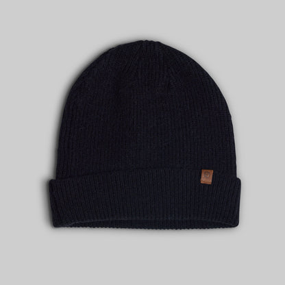 A dark navy blue ribbed knit beanie hat with a small brown leather brand patch on the cuff, shown against a solid gray background.