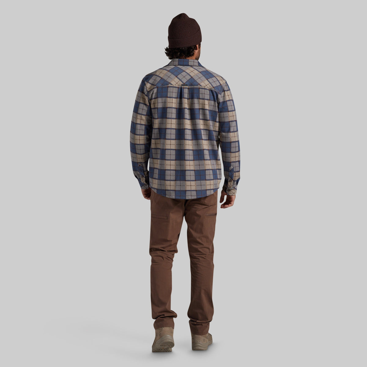 A man viewed from behind wearing a blue and beige plaid flannel shirt, brown pants, and a dark brown beanie against a solid gray background.