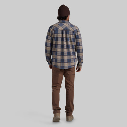 A man viewed from behind wearing a blue and beige plaid flannel shirt, brown pants, and a dark brown beanie against a solid gray background.