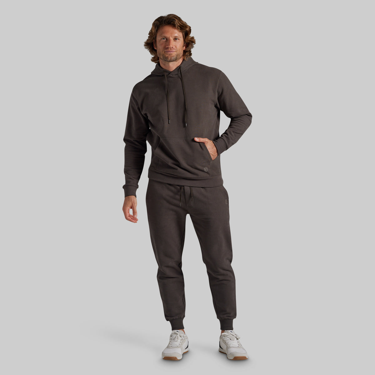 A man modeling a dark grey matching tracksuit, consisting of a pullover hoodie and jogger pants, paired with white sneakers, against a solid light gray background.