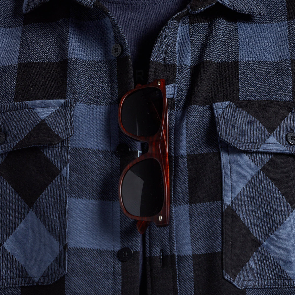 A close-up of a blue and black plaid flannel shirt with a pair of dark brown sunglasses tucked into the front pocket area.