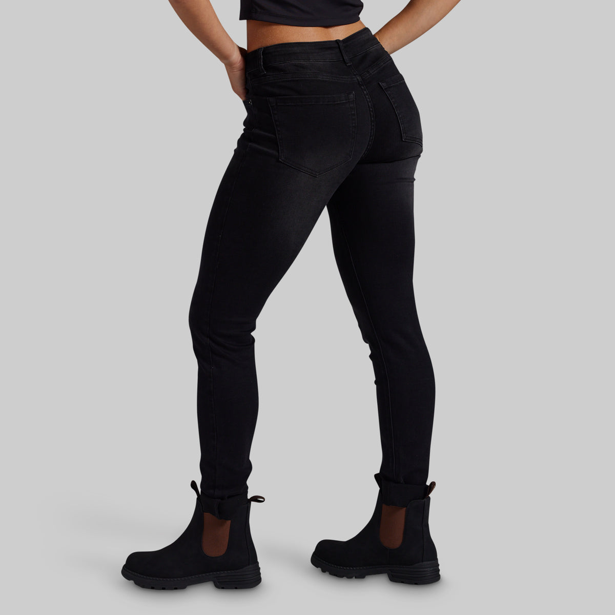 Rear three-quarter view of a model wearing black high-waisted skinny jeans and black Chelsea boots against a solid gray studio background.