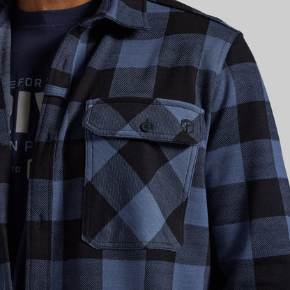 Close-up of a person wearing a blue and black buffalo plaid button-up shirt over a dark graphic t-shirt. The shirt features a chest pocket with a button and a small square logo patch.