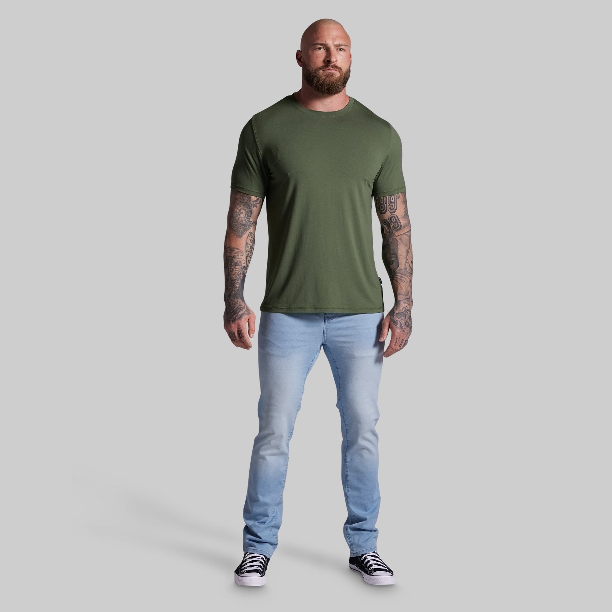 Man wearing an olive green t-shirt and light blue jeans on a gray background