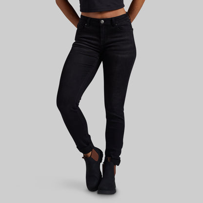A model wearing black slim-fit skinny jeans with rolled cuffs and black ankle boots, shown from the waist down against a solid gray background.
