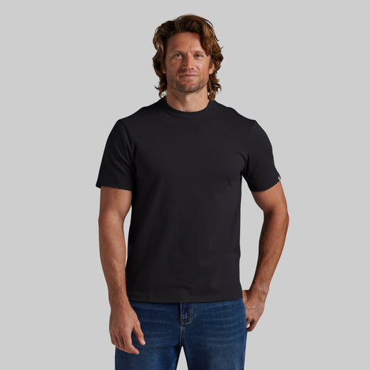 A man wearing a plain black short-sleeve crewneck t-shirt and blue jeans, standing against a solid light gray background.