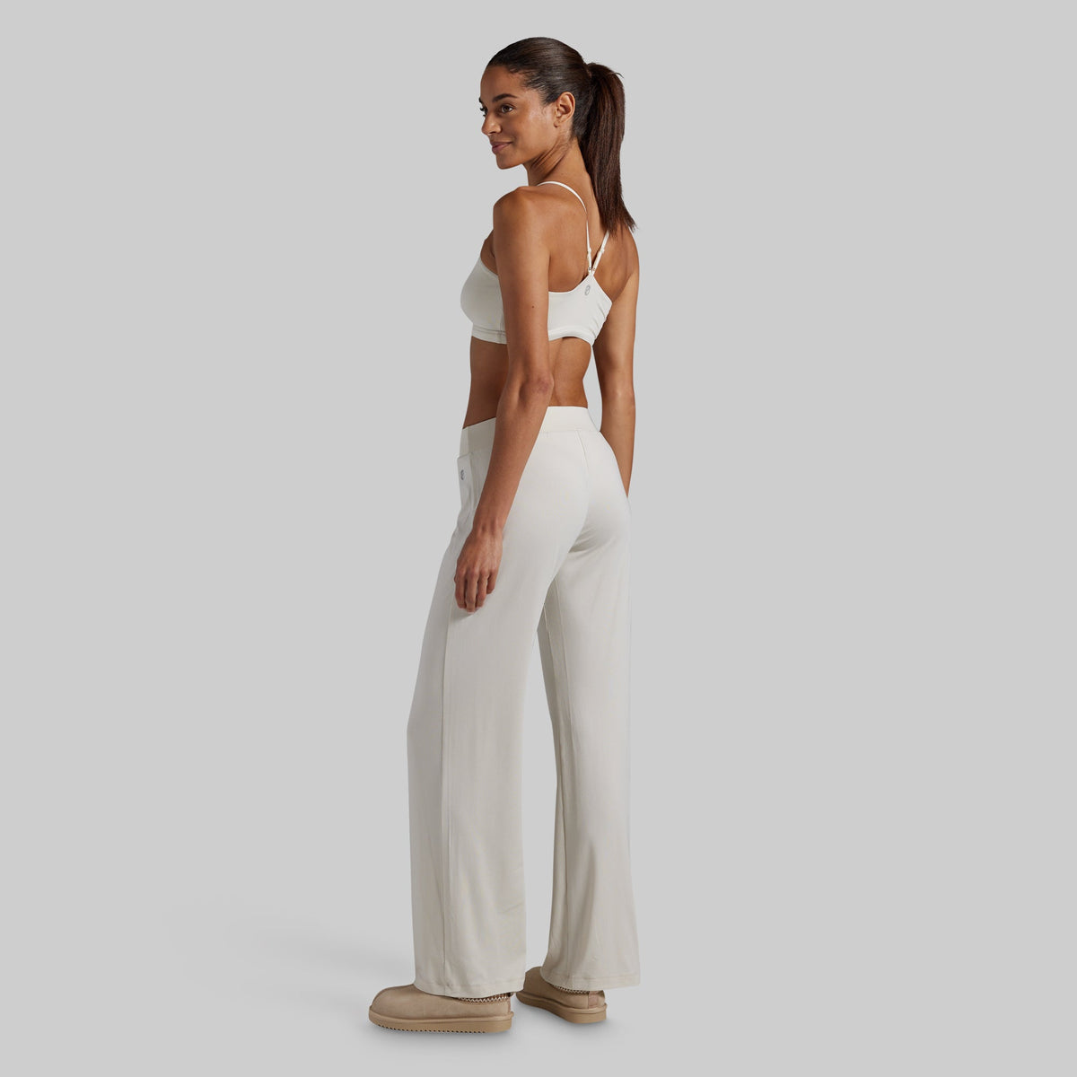 A woman stands in profile wearing a matching beige two-piece lounge set consisting of a sports bra and wide-leg pants, paired with beige slippers, against a solid light gray background.