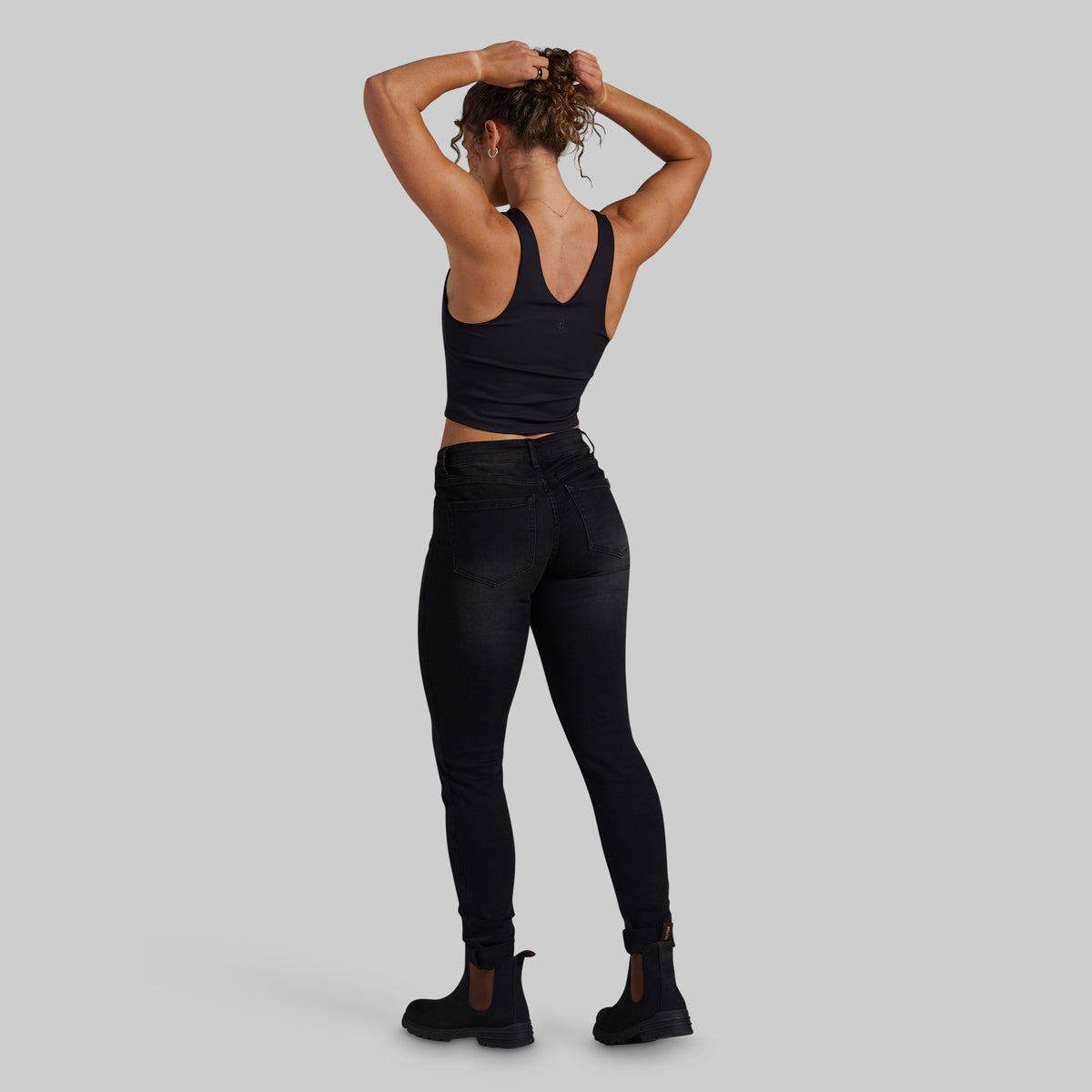 A woman shown from the back wearing a black racerback crop top, black high-waisted skinny jeans, and black ankle boots, standing against a solid light gray studio background.