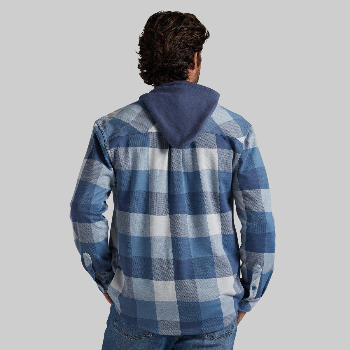 A man viewed from behind wearing a blue and white plaid hooded flannel with a solid navy blue hood, paired with blue jeans, against a solid gray background.