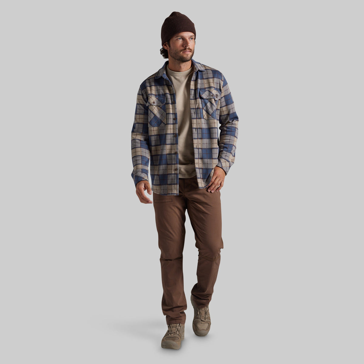 A man wearing a blue and beige plaid flannel shirt over a beige t-shirt, brown pants, and a dark brown beanie, walking against a solid gray background.
