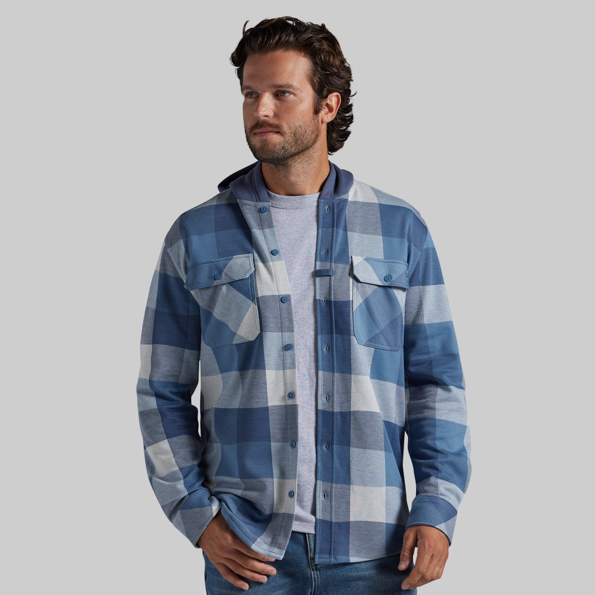 A man wearing a blue and white plaid hooded flannel shirt over a light gray t-shirt and jeans, standing against a solid gray background.