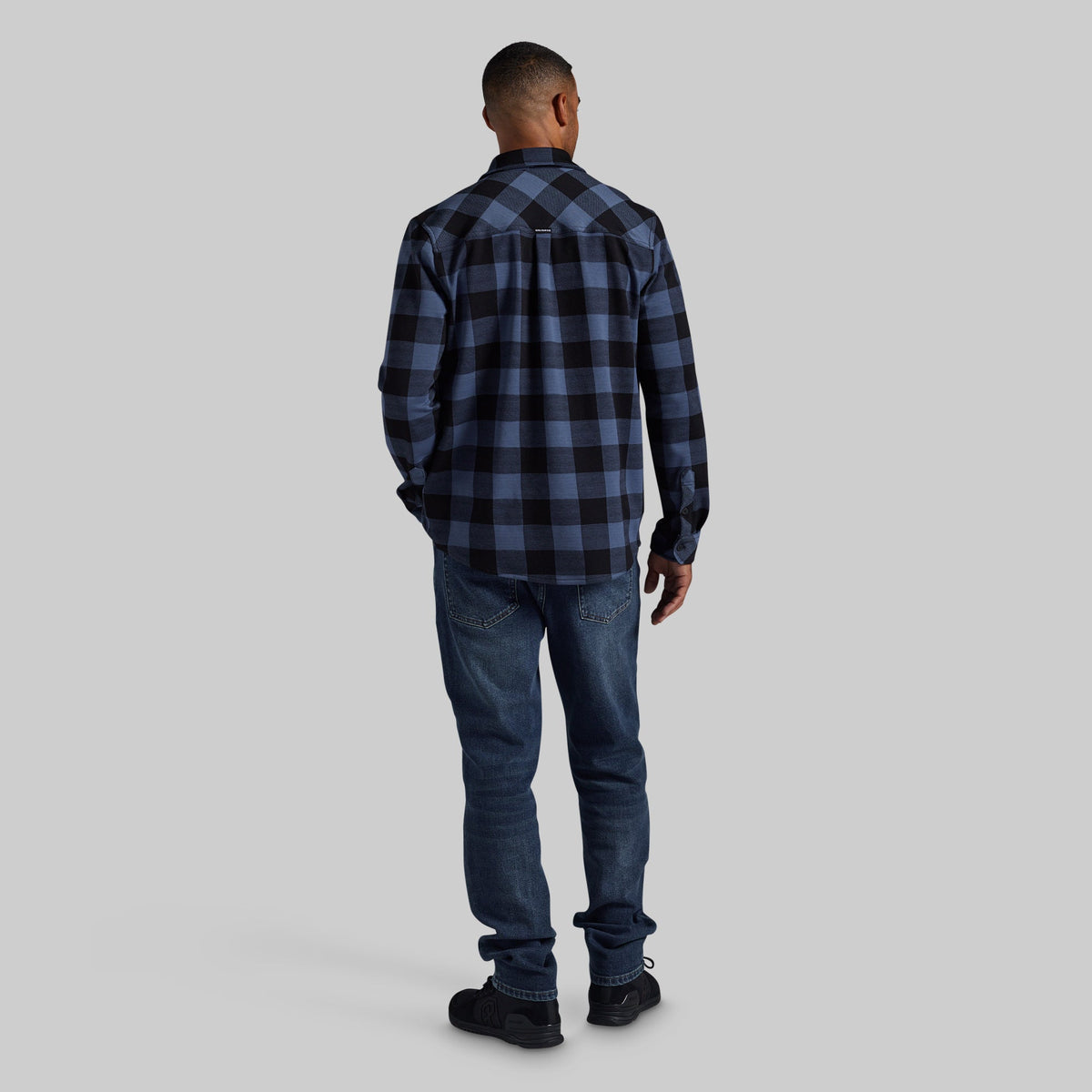 A man viewed from behind wearing a dark blue and black buffalo plaid flannel shirt, blue jeans, and black shoes, standing against a solid light gray background.