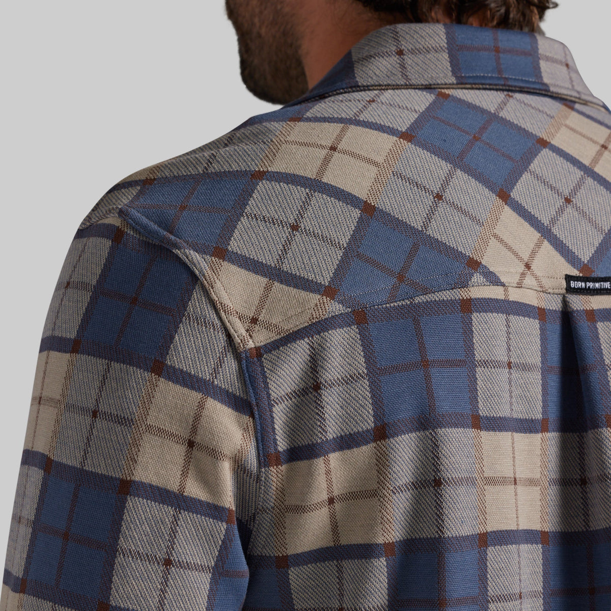 Close-up back view of a man wearing a plaid button-up shirt in blue, beige, and brown, with a small black brand tab on the upper right shoulder.