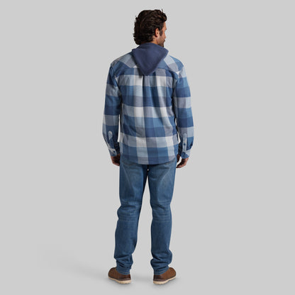 A man viewed from behind wearing a blue and white plaid hooded shirt, blue jeans, and brown shoes, standing against a solid gray studio background.