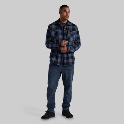 A man modeling a dark blue and black plaid flannel shirt with blue jeans and black sneakers, standing against a solid gray background.
