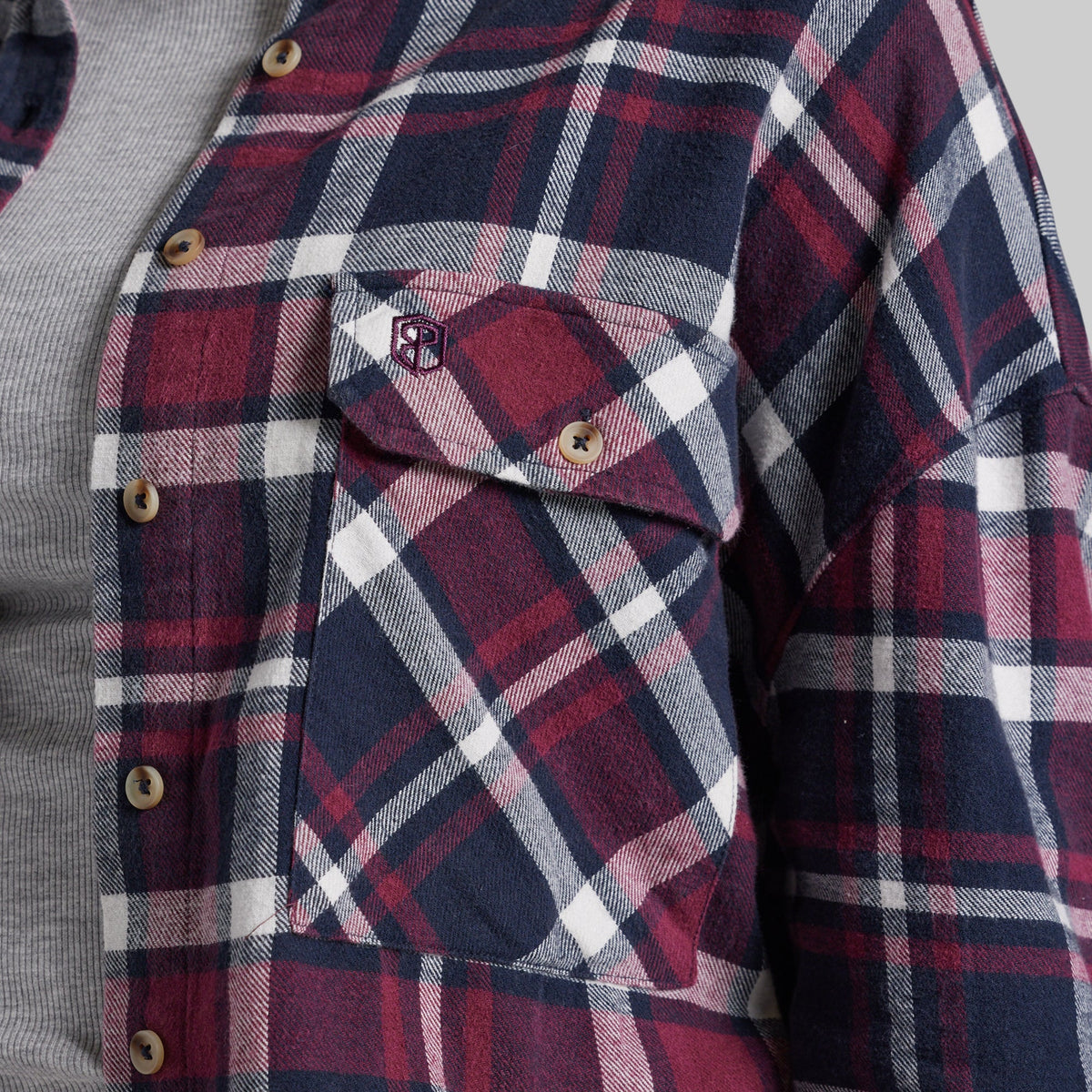 Close-up of a plaid button-up shirt with a chest pocket, featuring navy, burgundy, white, and gray stripes with a small embroidered logo on the pocket flap.