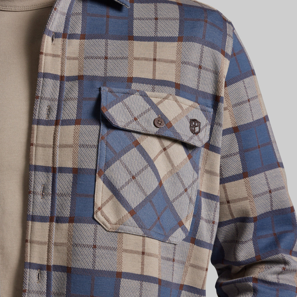 Close-up of a plaid button-up shirt with a chest pocket, featuring blue, beige, and brown tones, worn over a tan shirt against a light gray background.