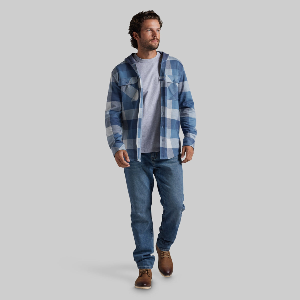 A man wearing a blue and white plaid hooded flannel shirt over a light gray t-shirt, blue jeans, and brown leather shoes, walking against a solid light gray background.