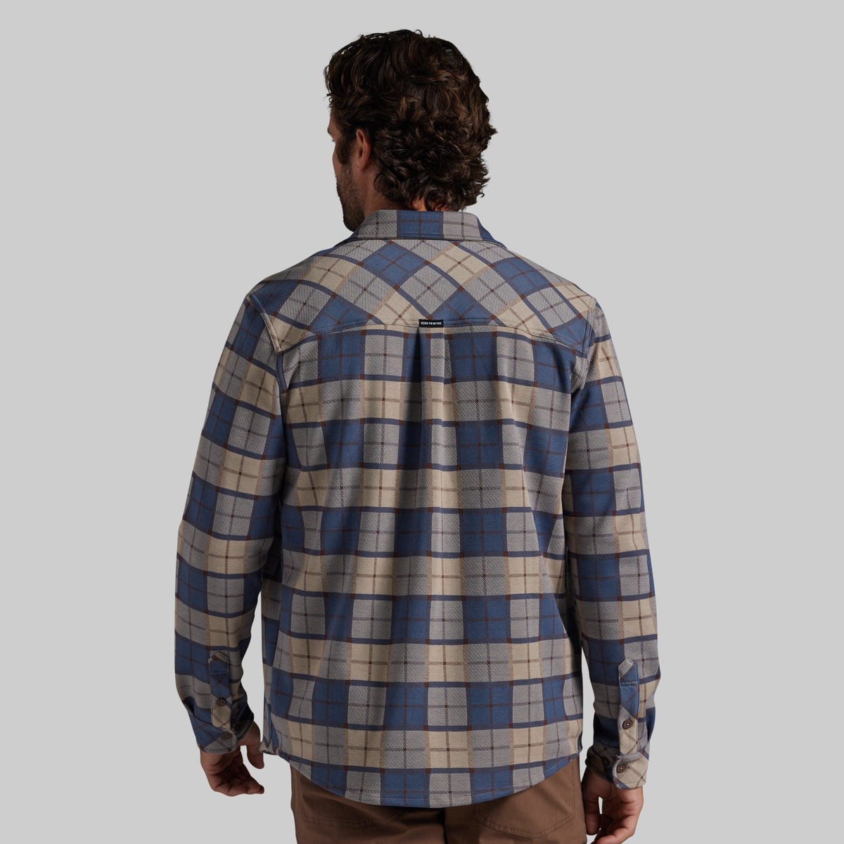 A man viewed from behind wearing a long-sleeve plaid button-up shirt in blue, beige, and gray, paired with brown pants, against a solid gray background.