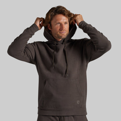A man wearing a dark brown pullover hoodie with drawstrings and a kangaroo pocket, adjusting his hood against a solid gray background.